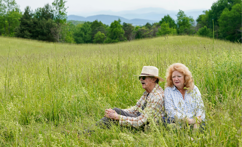 A man and woman sit in a field of tall grass. In the distance, trees and mountains are visible.