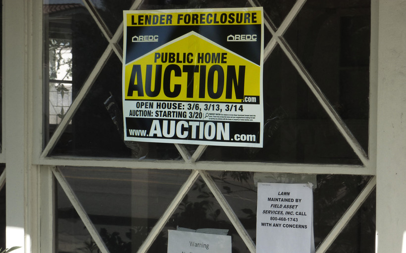 Window looking into a darkened foreclosed home. A yellow sign says "LENDER FORECLOSURE" and "PUBLIC HOME AUCTION." Smaller paper signs say "Warning No Trespassing" and note a lawn maintenance company.