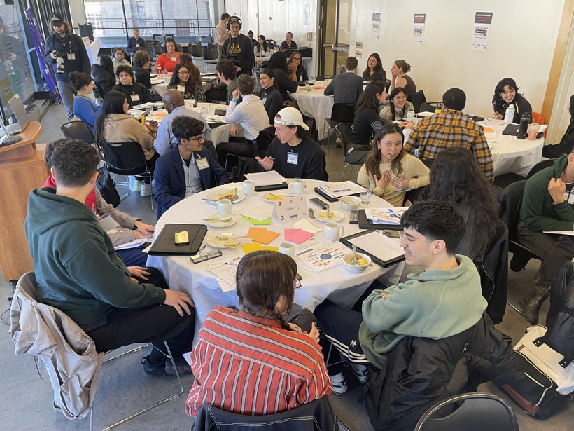 Groups of college-age people sit around round tables, engaged in discussions.