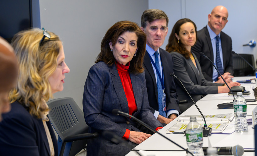 Governor Kathy Hochul, wearing a red turtleneck and blazer, sits at a table with other officials.