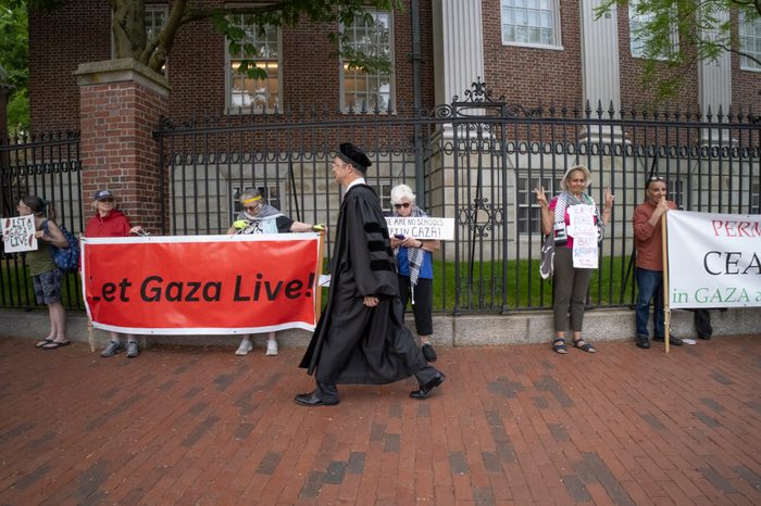 A male professor in a black academic robe and cap walks past a line of protesters outside Harvard holding signs and a red banner that reads "Let Gaza Live."