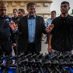 A man wearing glasses stands in front of a table full of rifles. He is surrounded by men, including some police officers.