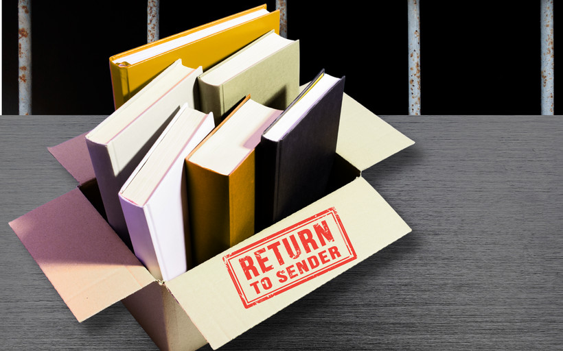 A box of books with a label reading "return to sender" sits on a metal table in front of prison bars.