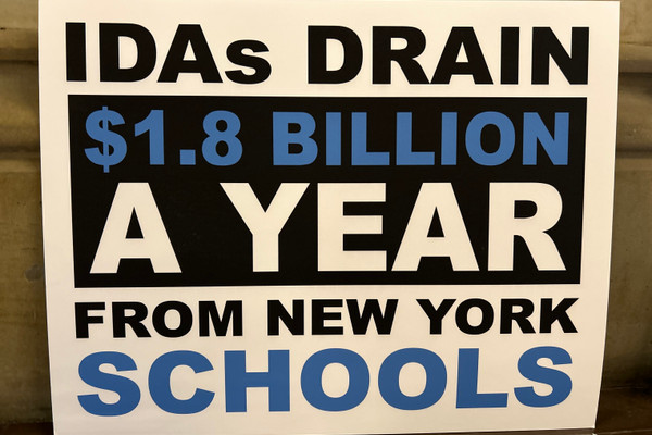 A sign reads "IDAs DRAIN $1.8 BILLION A YEAR FROM NEW YORK SCHOOLS."