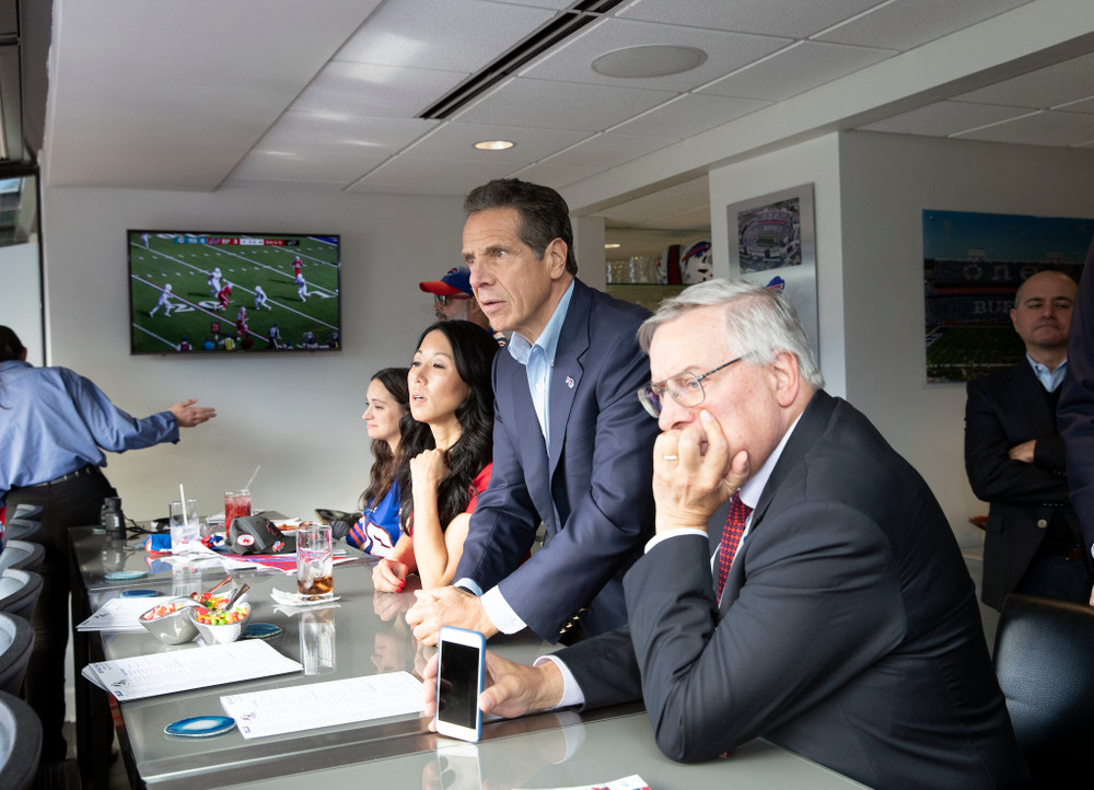 Several people, including former governor Andrew Cuomo of New York, stare out a well-lit window while a football game plays in the background.