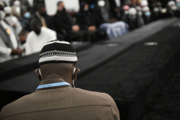 Close-up photo of a man from behind wearing a suit, kufi, and mask. In the background, many other people attending a funeral.