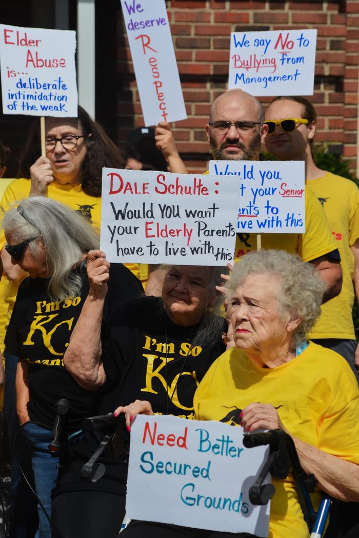 A groups of five or six elderly and younger people in yellow shirts hold signs decrying elder abuse and bullying, and demanding a more secure property.