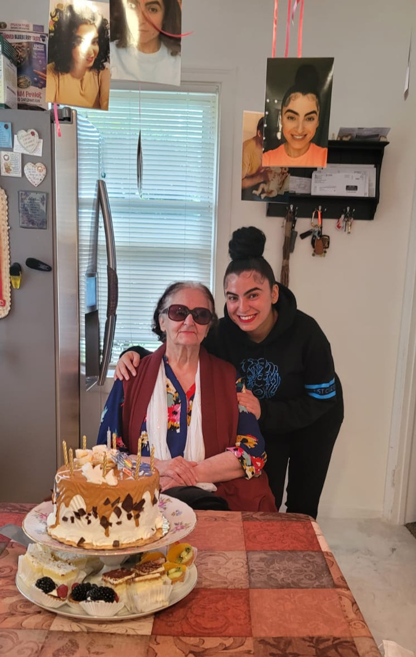 Saba Nakhai, a young woman, stands smiling behind and holds the arms of her older mother, who is seated and wearing sunglasses. The room seems celebratory; a cake is on the table in front of them, and pictures of Saba hang from the ceiling.