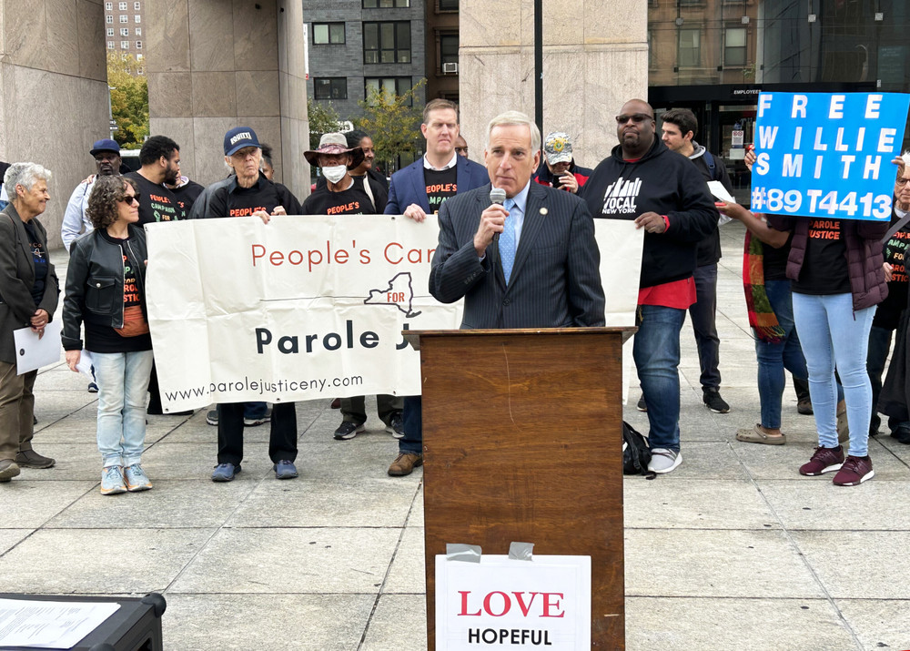 Assemblymember David Weprin stands at a podium in front of demonstrators holding signs advocating compassionate parole.