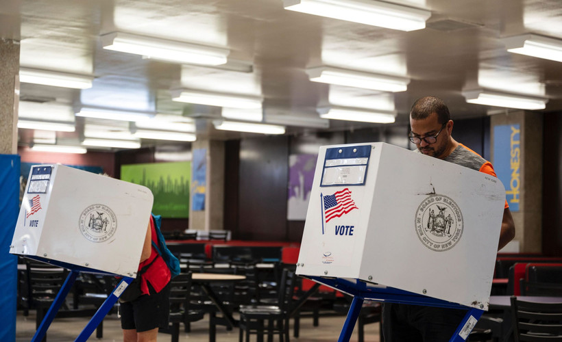 A man at a white voting booth with an american flag and a New York City insignia on it.