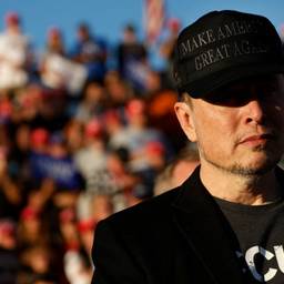 Elon Musk wears a MAGA hat, his eyes in shadow, with a blurry crowd behind him