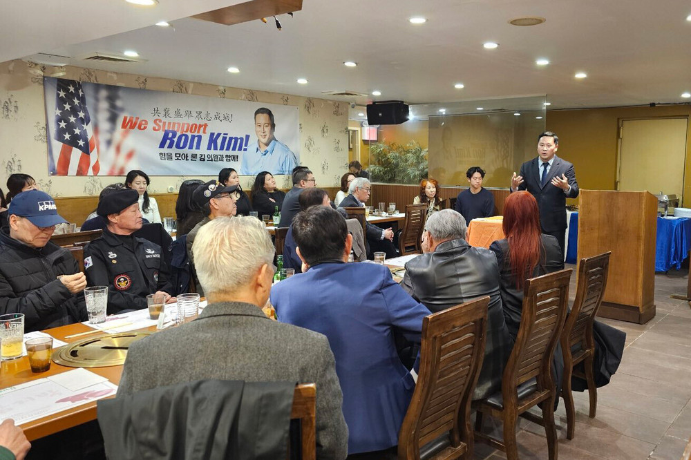 Assemblymember Ron Kim stands in a room full of people sitting at a long table. A poster on the wall reads "We Support Ron Kim!"