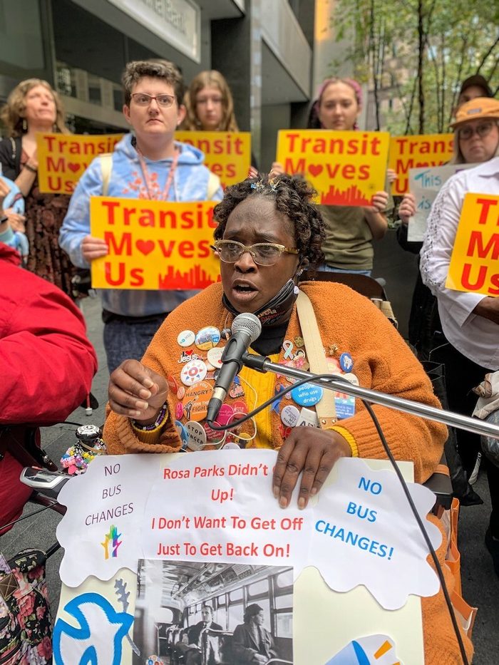 A Black woman speaks at a microphone holding a "Rosa Parks" sign. Behind her people hold yellow signs that say "Transit Moves Us" with a heart.