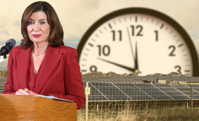 Governor Kathy Hochul stands at a podium in front of a field of solar panels. In the background, a large, looming clock ticks close to midnight.