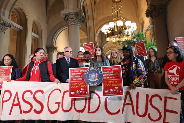 Activists and New York state Senator Julia Salazar rally to pass good cause eviction in Albany. They stand in the high-ceilinged halls of the Capitol with a white banner reading PASS GOOD CAUSE and various red signs.