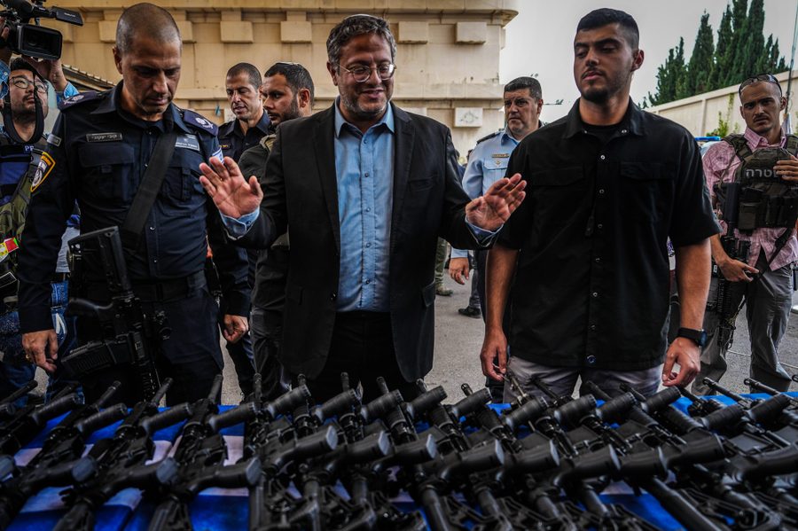 A man wearing glasses stands in front of a table full of rifles. He is surrounded by men, including some police officers.