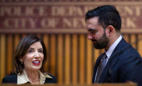 NYC Mayor Zohran Mamdani, in profile, looks at Governor Kathy Hochul, who looks outwards. The two stand close to each other behind a podium.