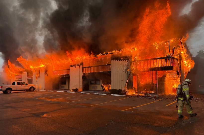 A motel is completely engulfed in flames, and black and gray smoke pours into the sky. A lone firefighter walks in the right side of the frame.