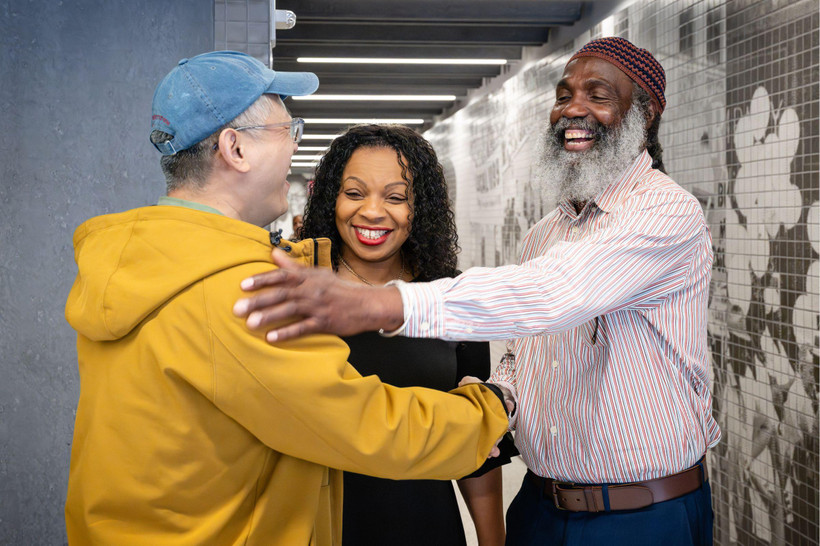 Three smiling people embrace collegially in an office hallway.