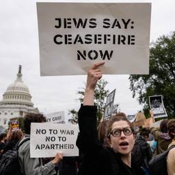 A person holds up a sign that reads "JEWS SAY: CEASEFIRE NOW". Behind them is another sign that reads "NO TO WAR NO TO APARTHEID". The U.S. Capitol Building is seen in the background, behind the crowd of protestors.