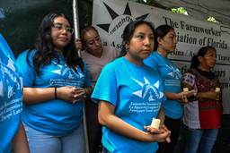 Three women hold candles; behind them, another weeps. They are in blue Farmworker Association t-shirts and traditional dress.