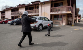 An adult and child wearing winter coats walk across a motel parking lot with three cars parked in it. The child is smiling and turns back to look at the adult, who is facing away from the camera.