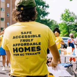 A masculine figure in a baseball cap stands with his back to the camera. His bright yellow shirt reads "Safe accessible truly affordable homes -- KC Tenants Union". Divers figures in yellow shirts stand int he background.