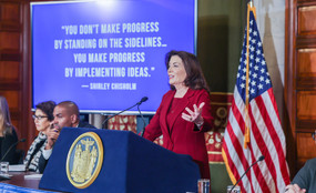 Governor Kathy Hochul speaks before a screen with a Shirley Chisolm quote, reading: "You don't make progress by standing on the sidelines... You make progress by implementing ideas."