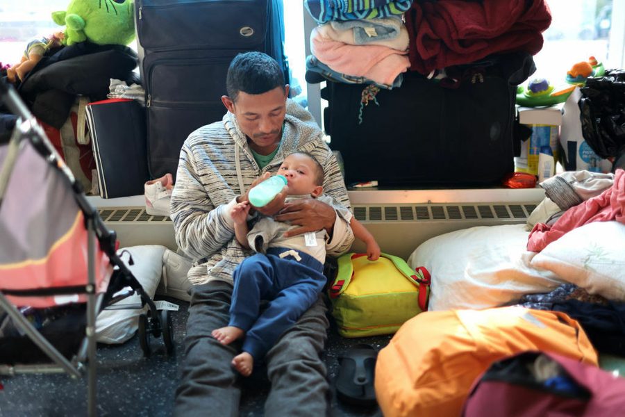 A man bottle-feeds an infant among strollers and sleeping bags.