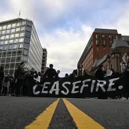 A group of protestors wearing all-black stand in the middle of a road. They hold a large black banner that says "CEASEFIRE" in white writing.