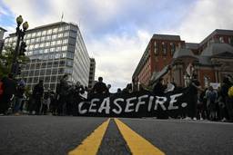 A group of protestors wearing all-black stand in the middle of a road. They hold a large black banner that says "CEASEFIRE" in white writing.