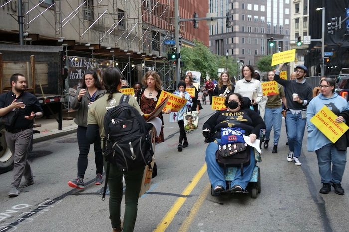 A person in a wheelchair leads a march down a busy pedestrian street