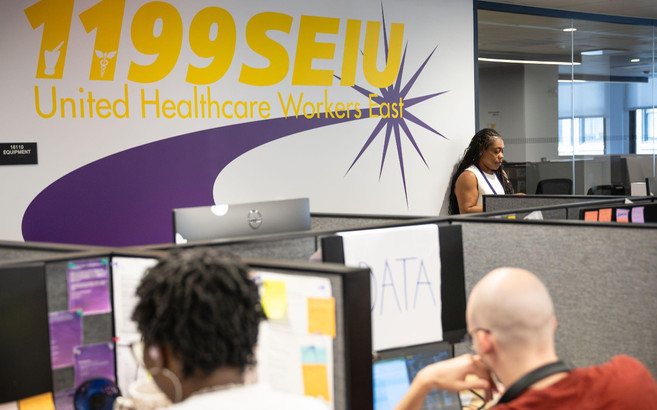 In the 1199 SEIU office, people work in at desks with a large 1199 SEIU: United Healthcare Workers East logo on the wall.