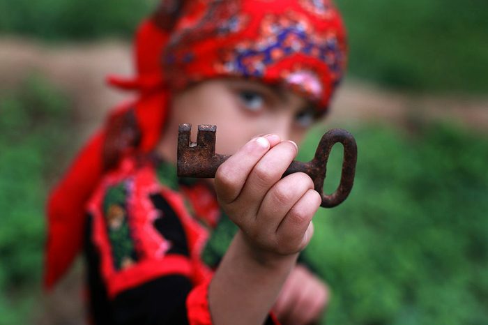 In the foreground is a child's hand holding a large, rusty key; behind it, slightly blurrt, his eyes look at the camera. He wears bright red festive clothing and scarf.
