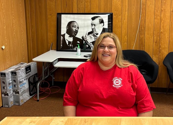 A woman in a red UAW shirt sits in an office in front of a framed portrait of MLK Jr and Walter Ruether