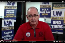 Screenshot of United Auto Workers President Shawn Fain in a livestream wearing a red UAW shirt, a UAW pin, and surrounded by UAW signs with demands like ?"End Tiers" and "COLA Fair Pay"