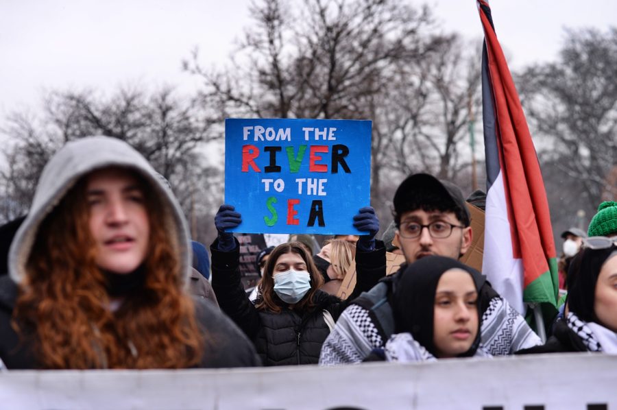A group of protestors stand in coats on DuSable Lake Shore Drive in Chicago. A protestor wearing a face mask and gloves, in the center of the image, holds a sign above their head, which reads "From the River to the Sea" in red, black, green and white letters. To their right, another protestor wearing a keffiyeh holds the Palestinian flag. Two other demonstrators in front of them are also wearing keffiyehs.
