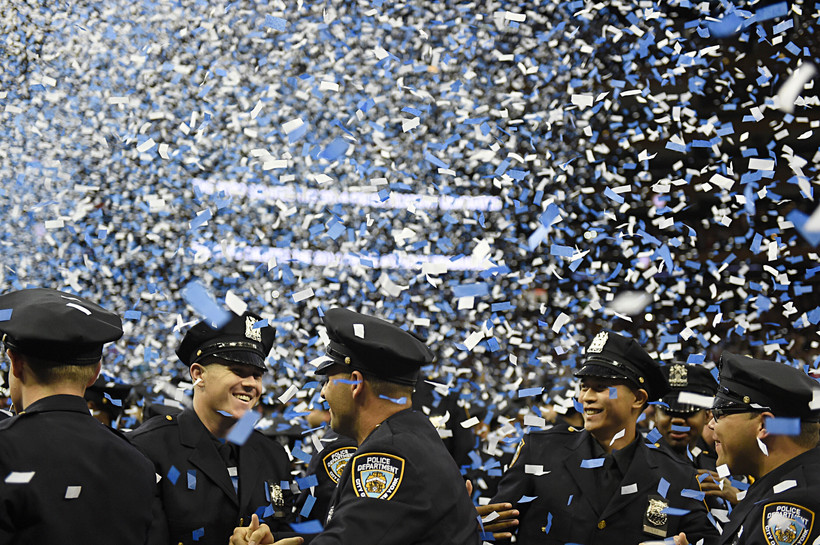 New NYPD officers smile amid blue and white confetti at police academy graduation ceremony.