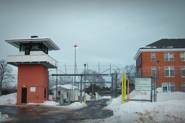 A photo of Marcy Correctional Facility shows a checkpoint for entering cars at a tall chainlink fence with barbed wire at the top. The area is snowy.