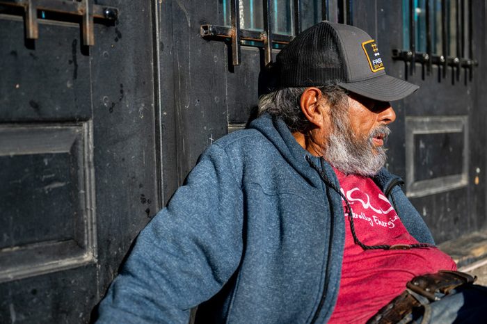 Closeup of a gray-bearded man in a baseball cap half-sitting half-lying against a wall.