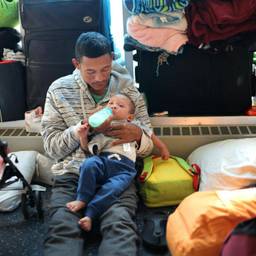 A man bottle-feeds an infant among strollers and sleeping bags.
