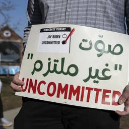 A sign in the foreground says "UNCOMMITTED" in red with Arabic text above, and a small image of a ballot with "uncommitted" checked and Biden unchecked. In the background is a woman in a hijab.