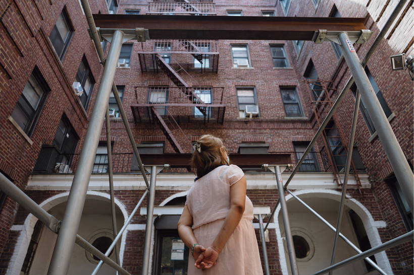 A woman stands outside a brick building, looking away from the camera, with her hands clasped behind her back.