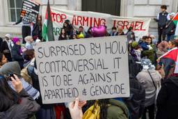 A group of people with banners and Palestinian flags stand outside a building in London. In the center of the photo, someone holds a sign that says, "Surprised by how controversial it is to be against apartheid and genocide."A banner in the background reads, in part, "Stop arming Israel."