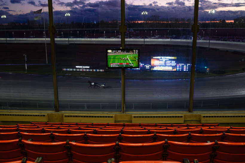 Rows of empty seats watch a horse racing track with a lone harness racer and horse.