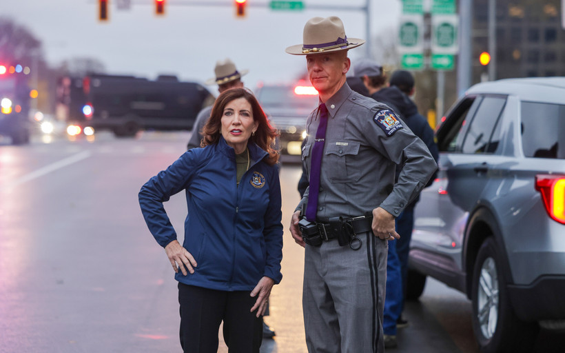 Governor Kathy Hochul stands with a hand on her hip next to a New York state trooper on a cloudy day.