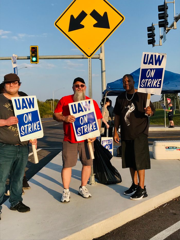 Three workers hold up UAW signs under a street sign in front of a traffic light.