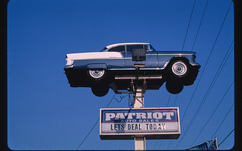 A blue car suspended on a post against a blue sky with a sign below reading "PATRIOT AUTO SALES: LETS DEAL TODAY"