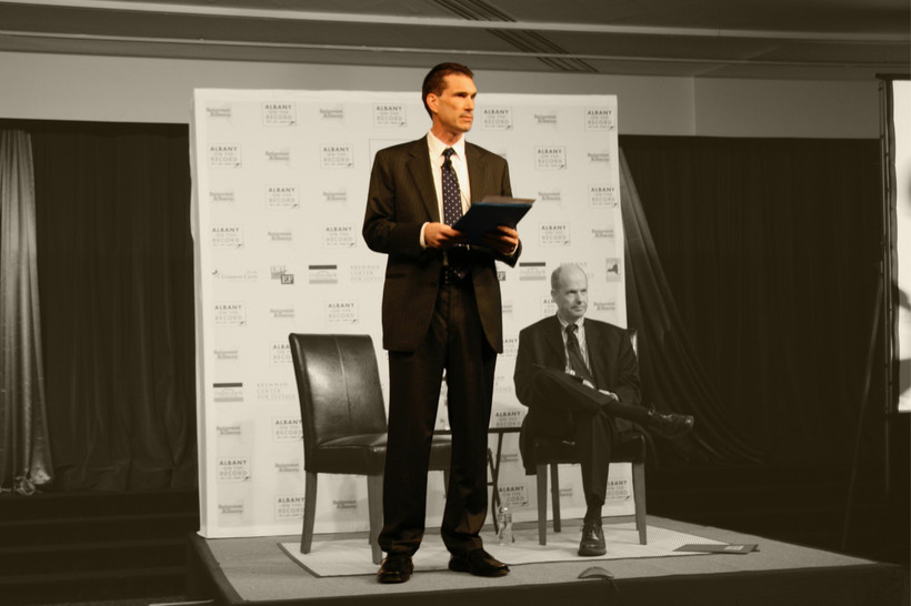 John Kaehny stands on a stage with a folder in his hands.
