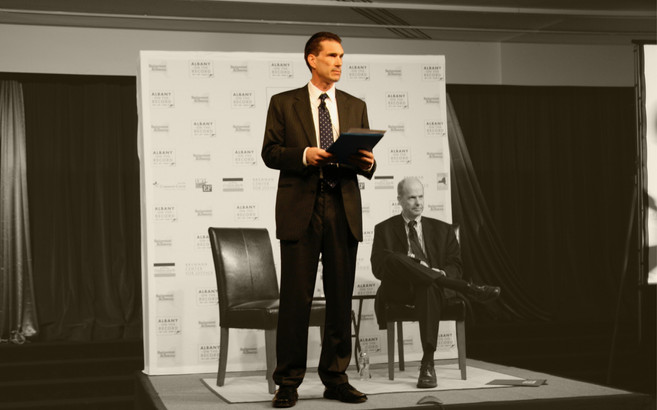 John Kaehny stands on a stage with a folder in his hands.